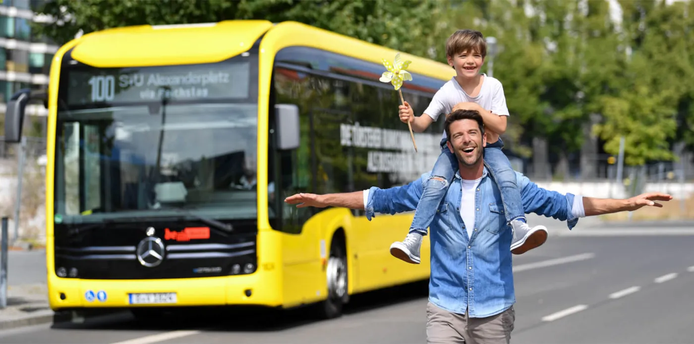 Ein Vater trägt seinen kleinen Sohn auf den Schultern. Im Hintergrund fährt ein Bus der BVG vorbei. 
