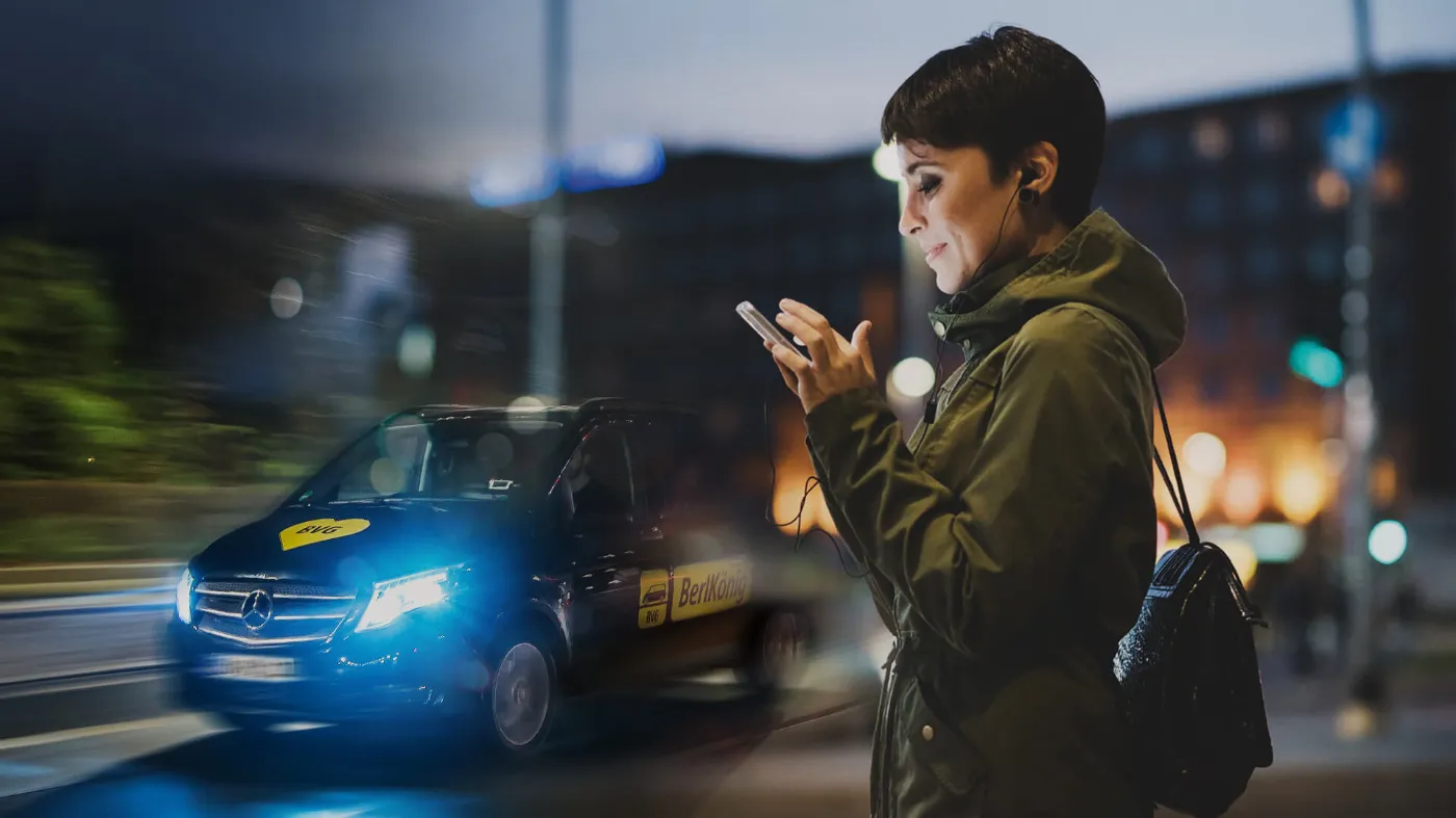 Eine junge Frau mit kurzen Haaren schaut auf ihr Smartphone. Im Hintergrund fährt ein BVG BerlKönig-Fahrzeug.