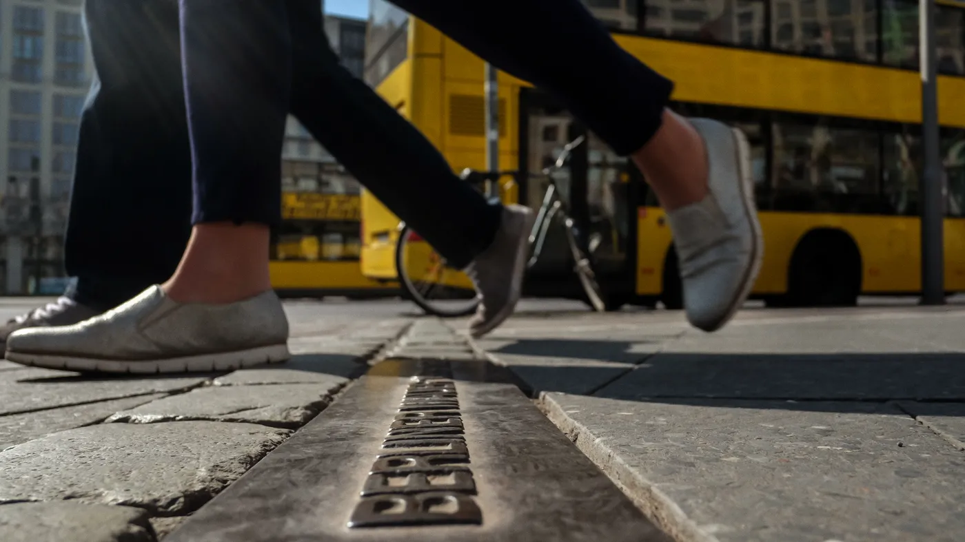 Close-up of 2 pairs of legs walking across a strip of the Berlin Wall. In the background a BVG bus.