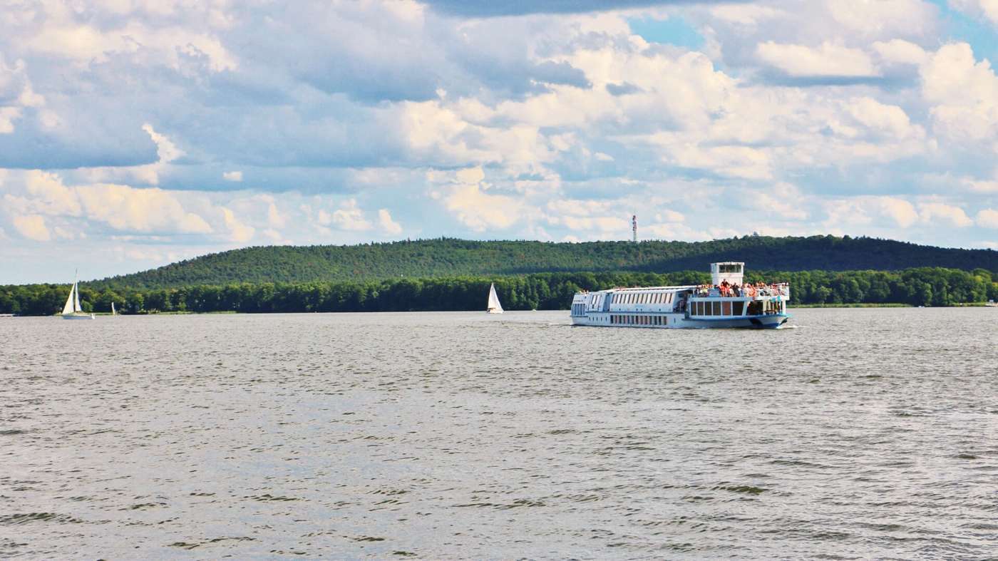 Zu sehen ist der Blick auf den Müggelsee. Auf dem See fährt ein Dampfer und zwei Segelboote.