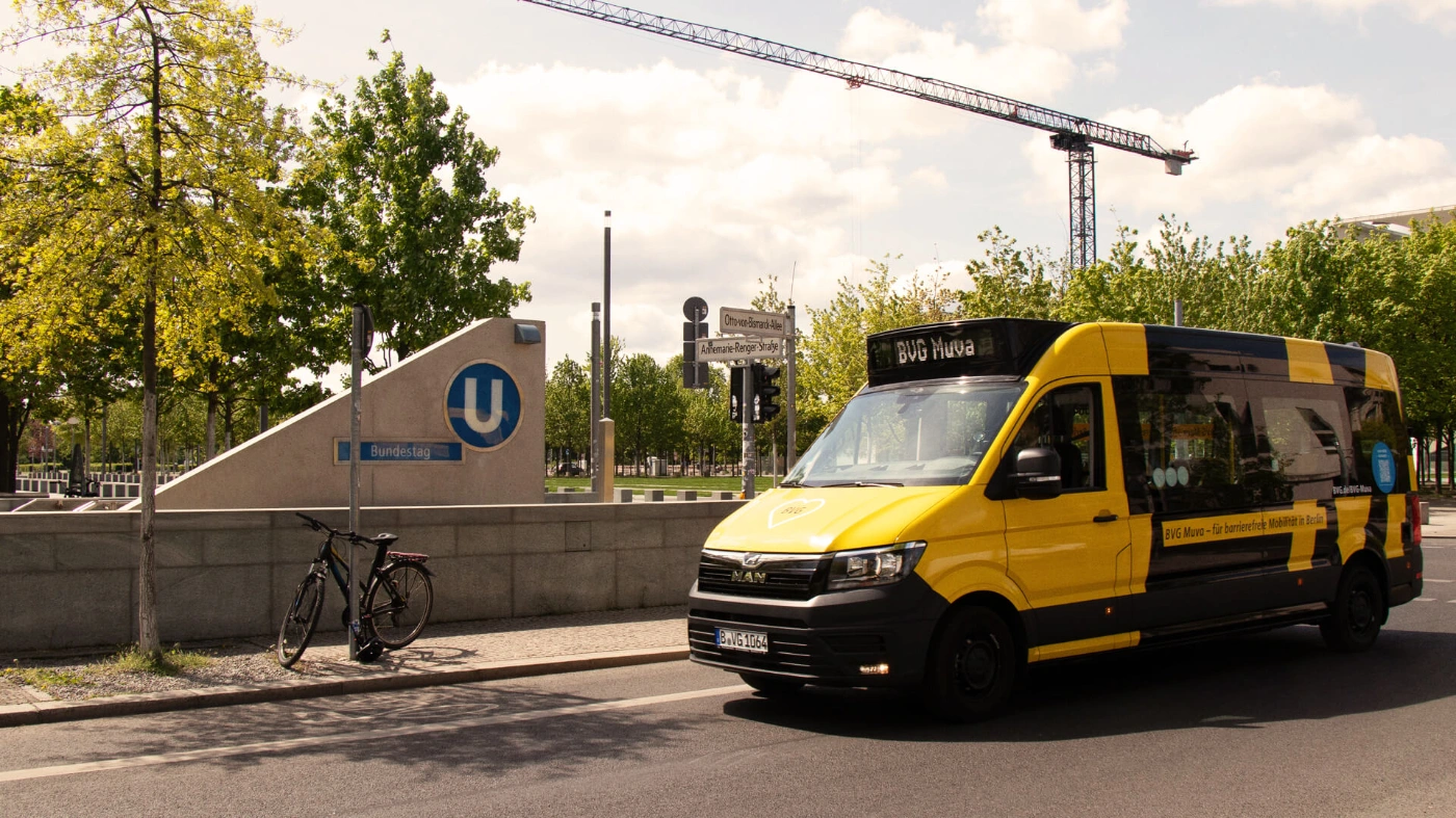 Ein Fahrzeug des BVG Muva fährt eine Straße entlang, direkt vor dem Eingang zum U-Bahnhof "Bundestag". Im Hintergrund ist ein großer Baukran zu sehen. "