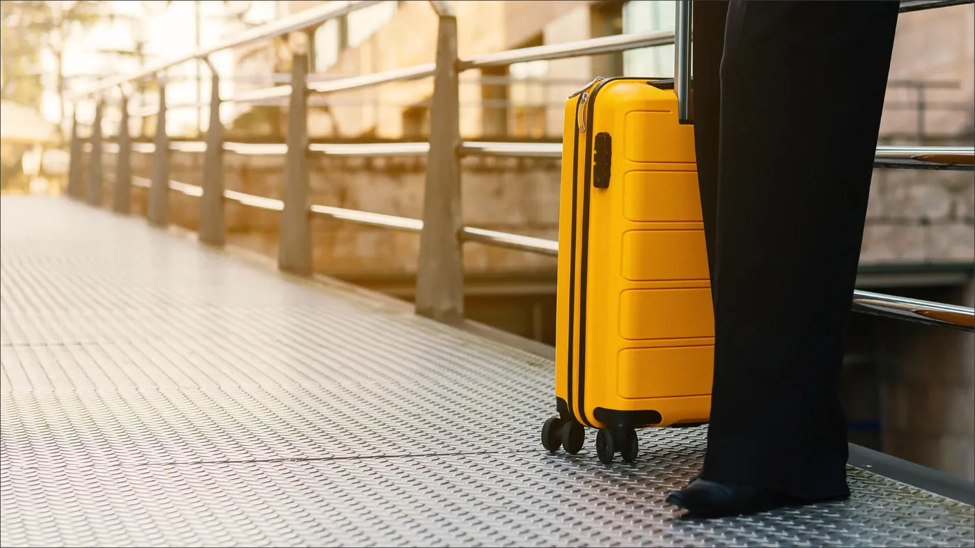 Nahaufnahme eines gelben Koffers auf dem Bahnsteig. Daneben steht eine Person in schwarzer Hose. Zu sehen sind nur die Beine der Person. 