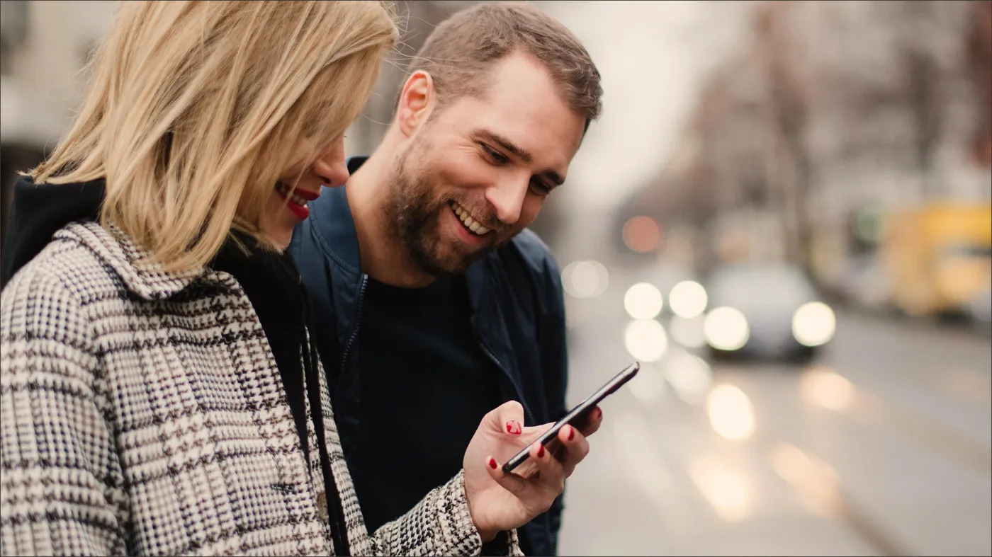 Ein Mann und eine Frau beugen sich lächelnd über ein Handy und schauen auf den Bildschirm. Die Frau hält das Handy in der Hand.