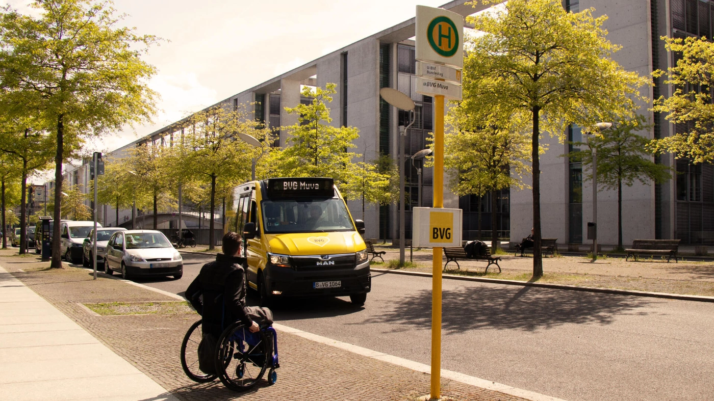 Eine Person im Rollstuhl wartet an einer Haltestelle der BVG. Auf die Haltestelle fährt ein BVG Muva zu. 