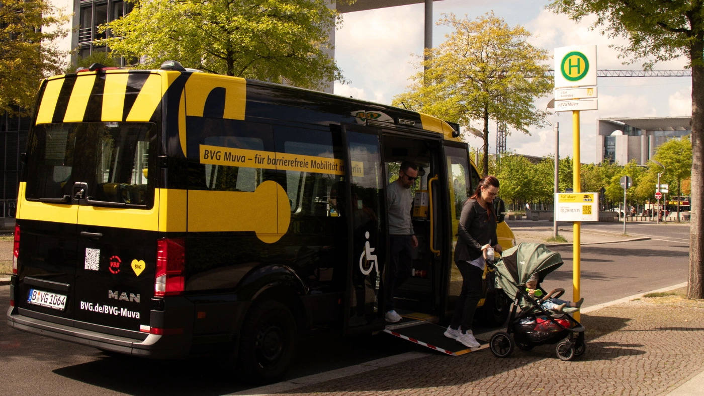 Ein Kleinbus des BVG Muva hält an einer Haltestelle. Ein junges Pärchen steigt aus, die Frau schiebt einen Kinderwagen aus dem Fahrzeug. 