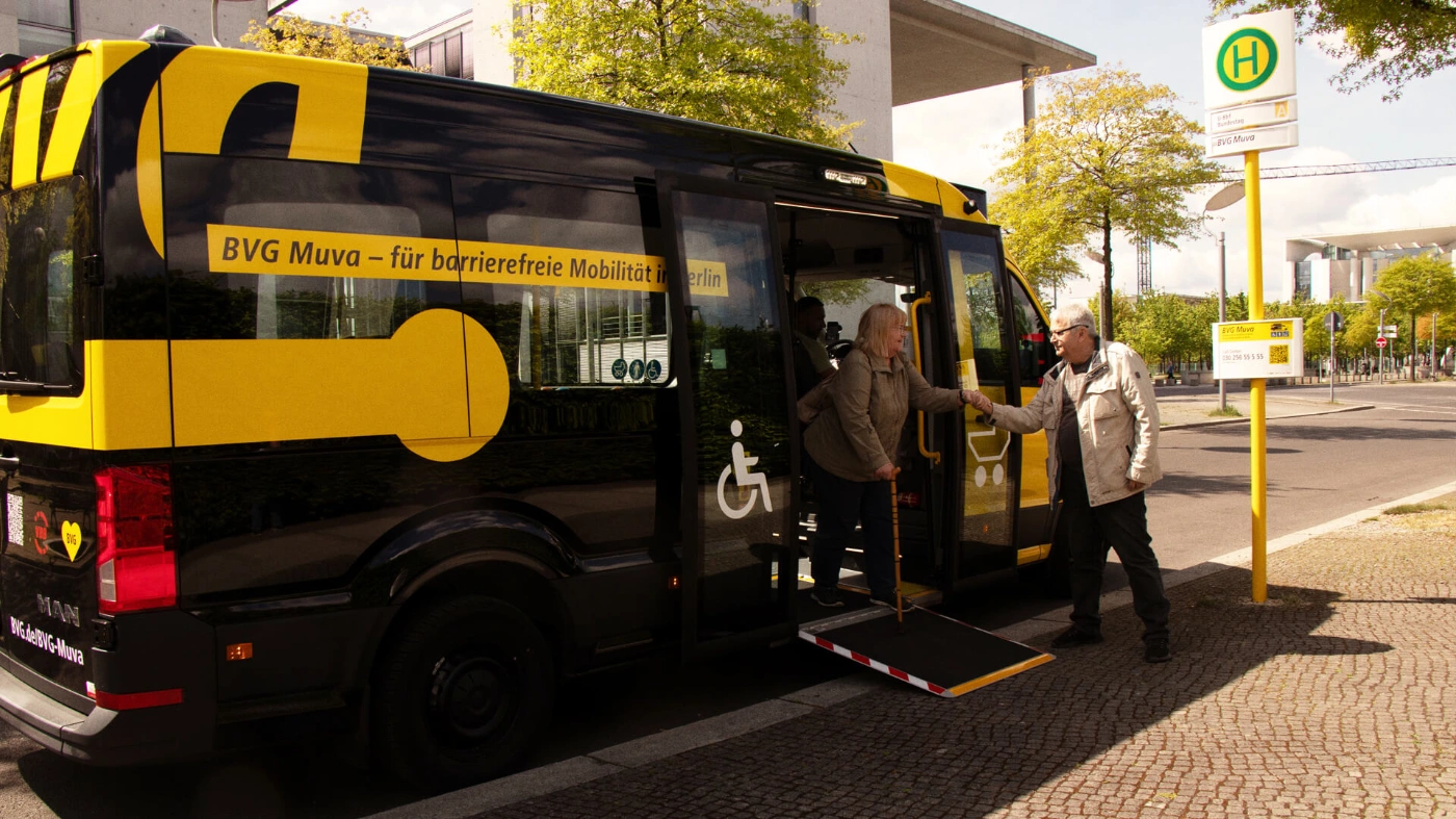 Ein Kleinbus des BVG Muva steht an einer Haltestelle. Der Fahrer hilft gerade einer Dame aus dem Fahrzeug, die eine Gehhilfe benutzt. 
