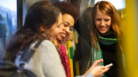 Three young women look at a cell phone while laughing on the train.