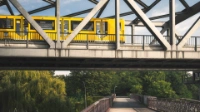 Über einen Park führt eine Brücke, darüber fährt eine gelbe U-Bahn der BVG. 