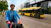 Eine Frau in einem blauen Blazer fährt Fahrrad, auf der gegenüberliegenden Straße fährt ein Bus der BVG vorbei. 