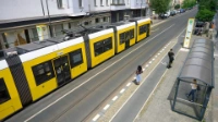 Eine Tram fährt in eine Haltstelle ein. Gegenüber warten Menschen. 