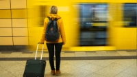 A woman with blond hair, yellow jacket and a suitcase is waiting for the train to arrive.