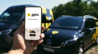 Eine Person hält ein Smartphone mit dem Logo des BVG Muva hoch, im Hintergrund sind zwei BVG Muva-Fahrzeuge zu sehen. 