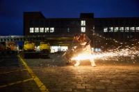 Ein Auszubildender der BVG arbeitet abends auf dem Betriebsgelände mit einer Trennschleifmaschine, während Funken fliegen – im Hintergrund gelbes Gleisbaugerät und der BVG-Ausbildungs-Campus.