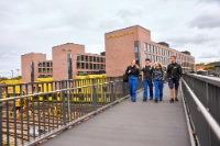 Vier Auszubildende der BVG gehen lachend über eine Fußgängerbrücke mit Blick auf den BVG-Ausbildungs-Campus in Berlin; im Hintergrund sind gelbe Arbeitsfahrzeuge und Gleisanlagen zu sehen.