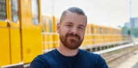 Auszubildender Florian steht auf einem BVG-Betriebsgelände vor den Gleisen. Im Hintergrund ist eine U-Bahn zu sehen.