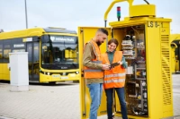 Ein Ingenieur und eine Ingenieurin stehen vor einer Ladesäule. Im Hintergrund steht ein E-Bus