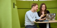 Zwei Personen sitzen auf einer grünen gepolsterten Bank und schauen auf einen Laptop.