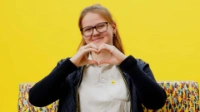 Eine lächelnde BVG-Busfahrerin sitzt auf einer Bank mit buntem Diversity-Muster vor gelbem Hintergrund und formt mit beiden Händen ein Herz.