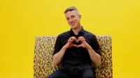 Ein lächelnder Ingenieur der BVG sitzt auf einer Bank mit buntem Diversity-Muster vor gelbem Hintergrund und formt mit beiden Händen ein Herz.