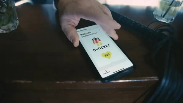 In Großaufnahme ist die Hand einer Person zu sehen, die ein Handy auf den Tresen legt. Auf dem Bildschirm ist das Deutschlandticket zu sehen. 