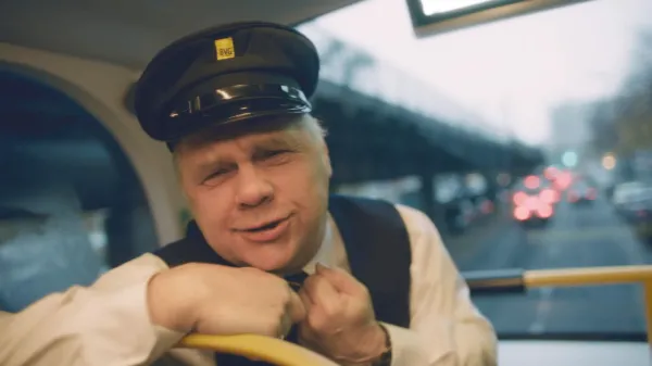 Ein Busfahrer der BVG dreht sich von seinem Platz um, um in die Kamera zu sprechen und gestikuliert dabei lächelnd. 
