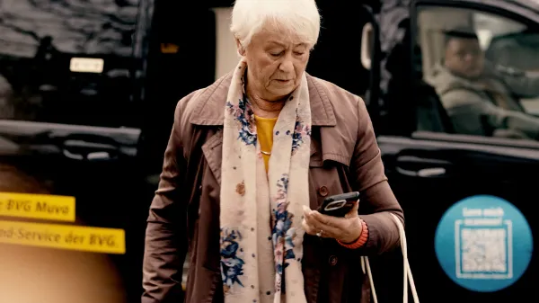 Eine ältere Frau schaut auf ein Handy in ihrer Hand. Im Hintergrund ein Muva-Fahrzeug.